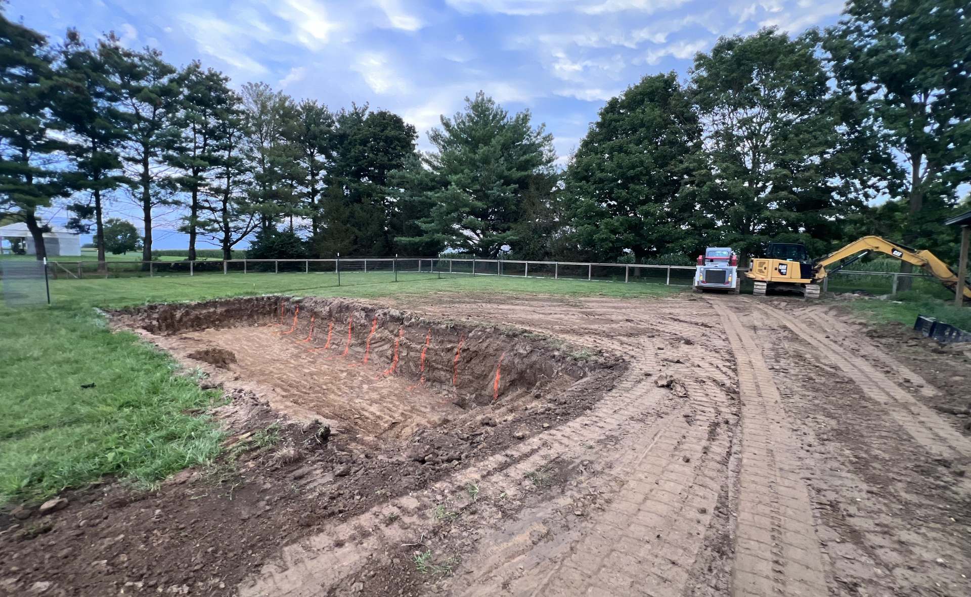 A rectangular excavation opened in a rural yard, with an excavator and track loader at the fence