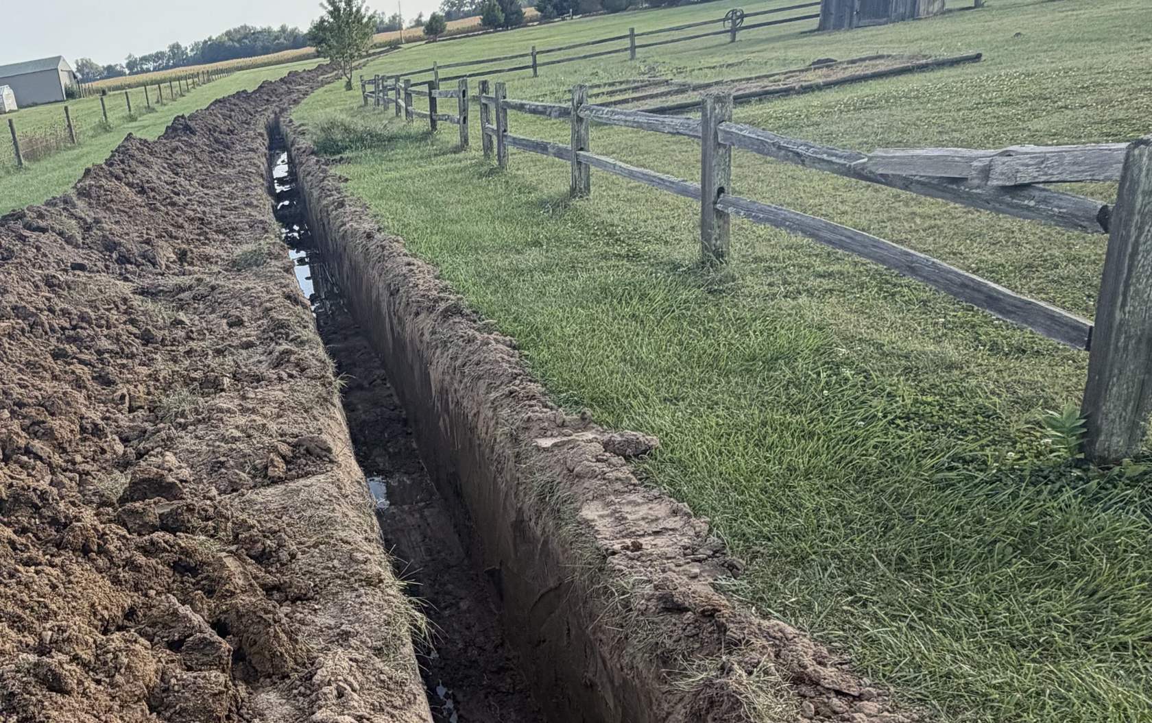 A long open trench cut along a split rail fence on a rural property